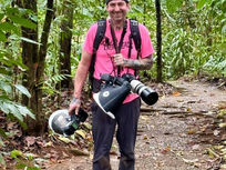 Man in a pink shirt and cap stands on a forest trail holding cameras. Lush green foliage surrounds him. He looks happy and enthusiastic.