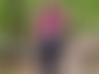 Man in a pink shirt and cap stands on a forest trail holding cameras. Lush green foliage surrounds him. He looks happy and enthusiastic.