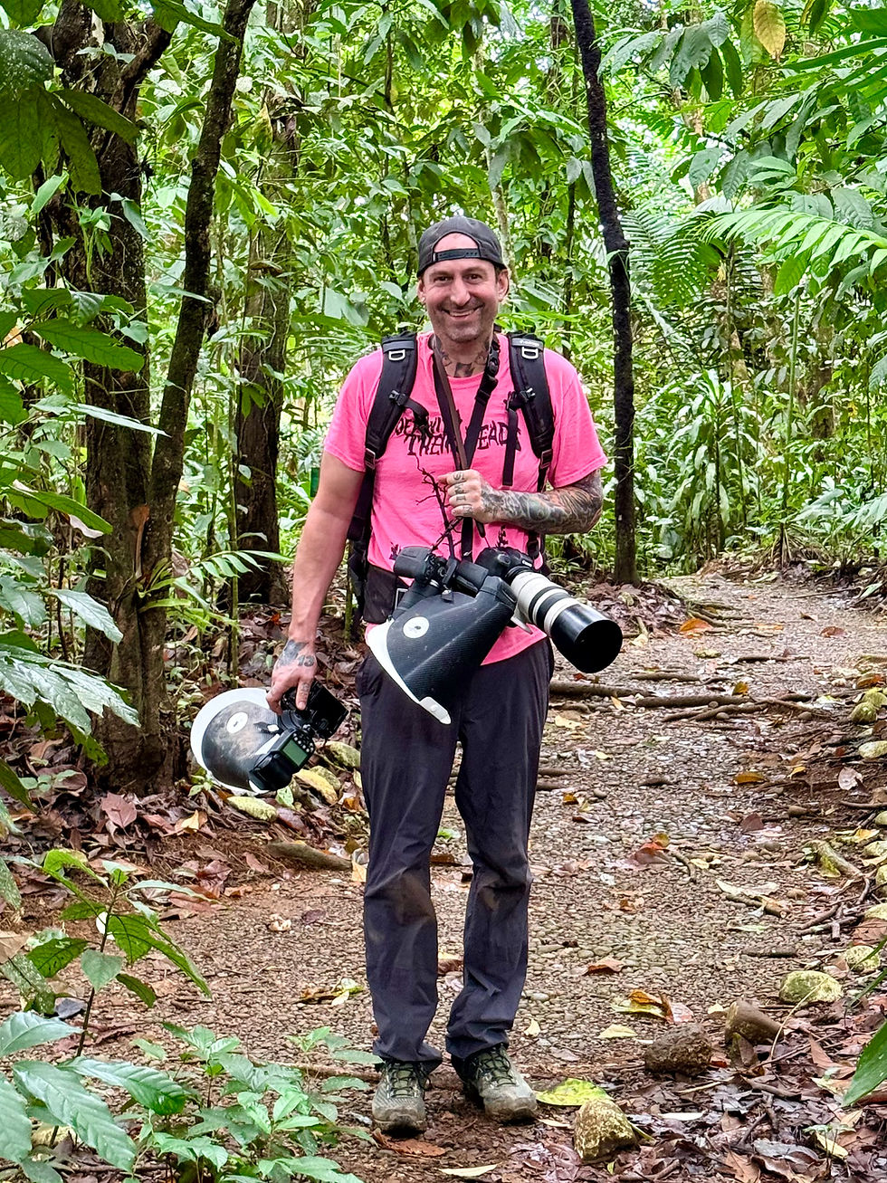 Man in a pink shirt and cap stands on a forest trail holding cameras. Lush green foliage surrounds him. He looks happy and enthusiastic.