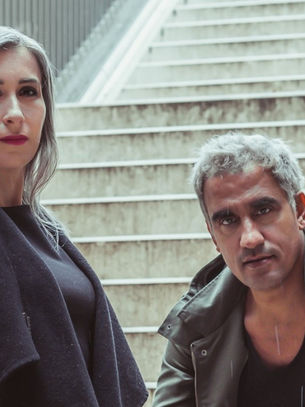 Two people with gray hair, one standing and one crouching, on a stone stairway. Overcast mood, neutral tones, no text visible.