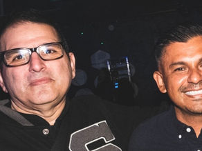 Two smiling men pose arm-in-arm in a dark indoor setting, one wearing glasses.