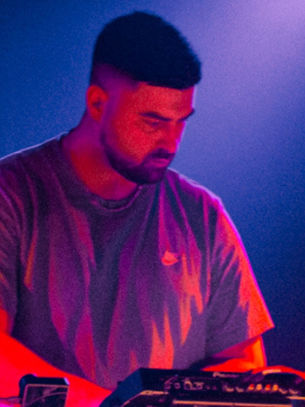 A person in a dimly lit setting operates a sound mixer, illuminated by purple and blue lights, creating a focused and creative mood.