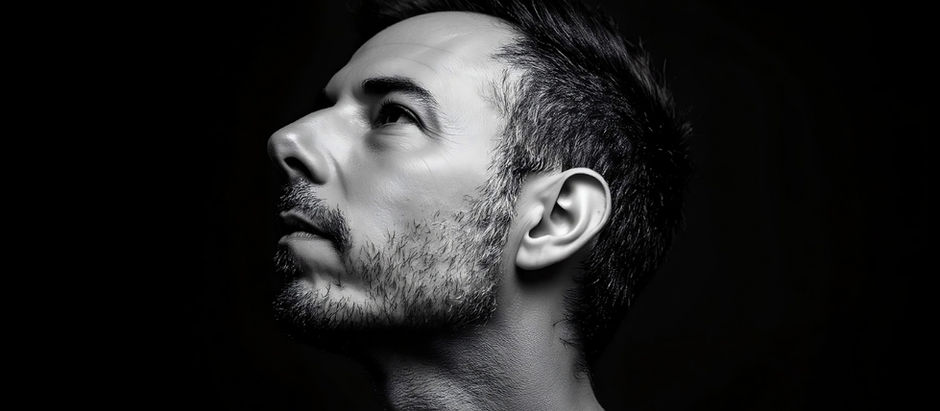 Side profile of a man looking up, in black and white. His expression is contemplative against a dark background, highlighting facial details.