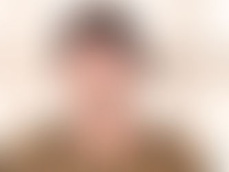 Man in a colorful sweater stands against a textured glass window, with neutral expression. The sweater is olive with orange and red stripes.