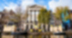 Canal-side row of historic European buildings with trees, boats, and pedestrians under a bright blue sky.