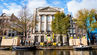 Canal-side row of historic European buildings with trees, boats, and pedestrians under a bright blue sky.
