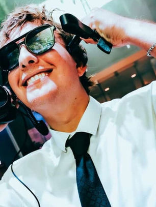 DJ in sunglasses and white shirt smiling, holding headphones to ears. Bright stage lights, DJ mixer visible. Energetic party vibe.
