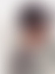 Man wearing a black hat and sunglasses stands against a plain white wall. He has a trimmed beard and wears a grey jacket and white shirt.