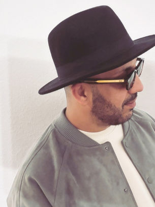 Man wearing a black hat and sunglasses stands against a plain white wall. He has a trimmed beard and wears a grey jacket and white shirt.