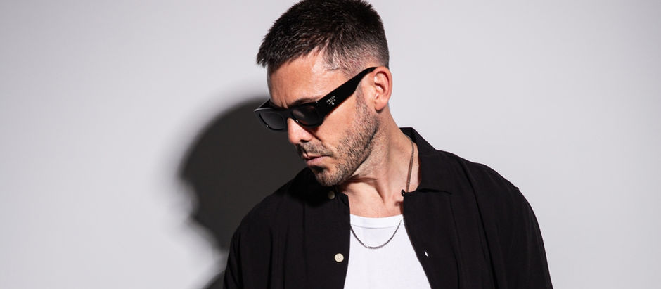 Man in black sunglasses and shirt with a white tee, looking down. Shadow on a plain gray background creates a moody effect.