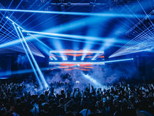 Concert crowd with hands raised under blue laser lights and smoke, creating an energetic atmosphere. Bright stage with red and blue visuals.