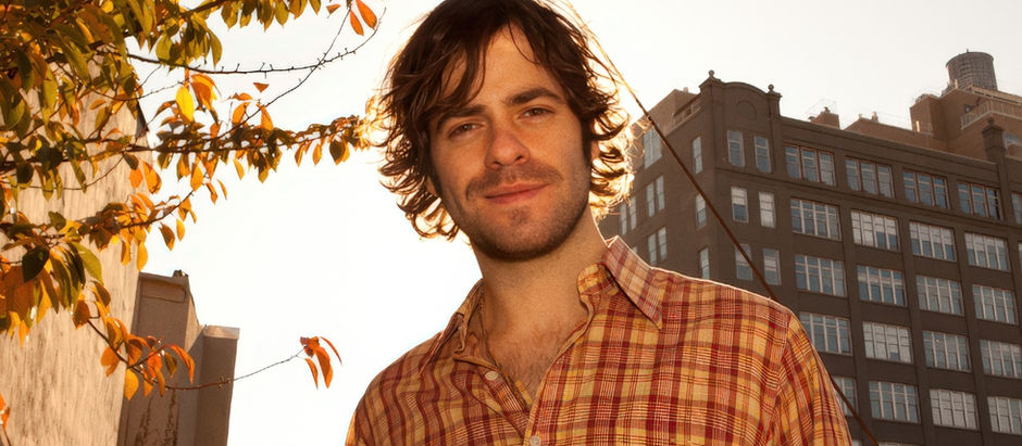 Smiling man in a red plaid shirt stands outdoors by autumn leaves, backlit against city buildings.