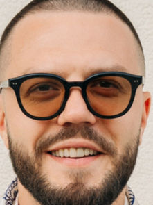 Man with a buzz cut and beard smiling, wearing dark sunglasses. Background is a plain, textured white wall. Mood is casual and upbeat.