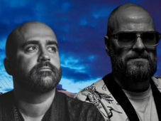 Two men with serious expressions, one wearing sunglasses, against a vivid blue sky with architectural structures. Black and white attire.