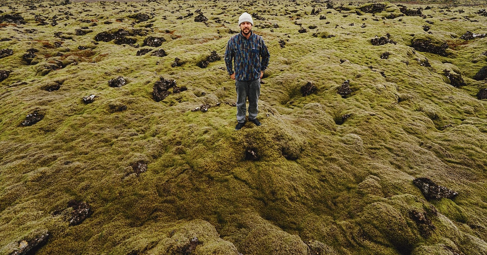 Man in plaid shirt and white hat stands on a mossy terrain with black rocks. Overcast sky creates a serene, natural setting.