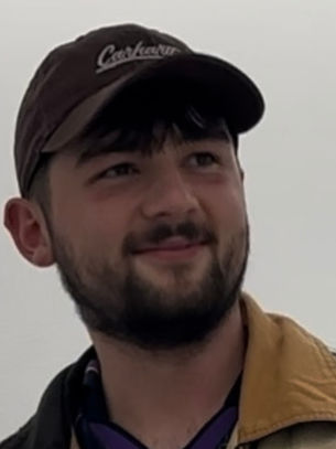 Young man with a beard and a cap smiles subtly. He wears a tan jacket with a white background. The cap has indistinct text. Mood is relaxed.