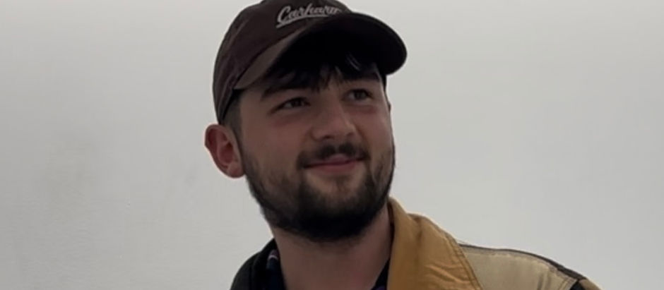 Young man with a beard and a cap smiles subtly. He wears a tan jacket with a white background. The cap has indistinct text. Mood is relaxed.