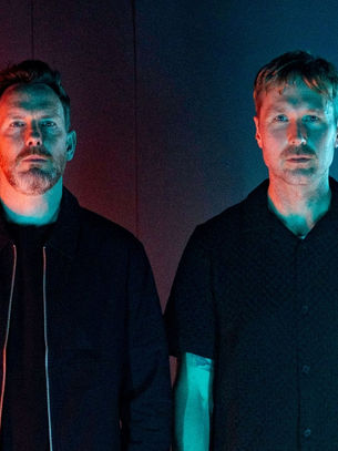 Two men stand side by side in a dimly lit room. One is under red light, the other under blue, creating a moody, mysterious vibe.