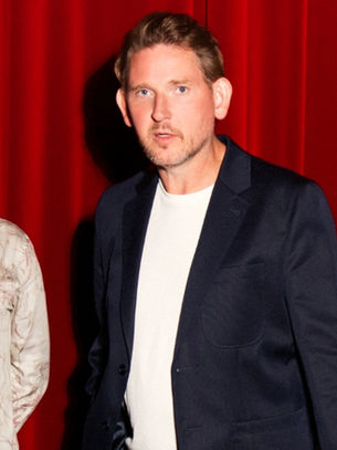 Three men pose seriously in front of a red curtain, wearing a patterned shirt, dark blazer, and light blazer.