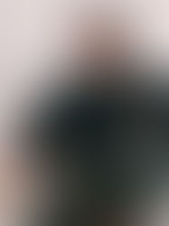 Man sitting in an orange chair, wearing a black jacket, camo pants, and sneakers. White background, relaxed expression.