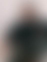 Man sitting in an orange chair, wearing a black jacket, camo pants, and sneakers. White background, relaxed expression.