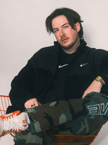 Man sitting in an orange chair, wearing a black jacket, camo pants, and sneakers. White background, relaxed expression.