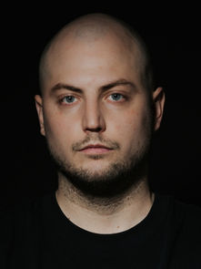 A bald man with a serious expression is photographed against a dark background, wearing a black shirt. The mood is somber and intense.