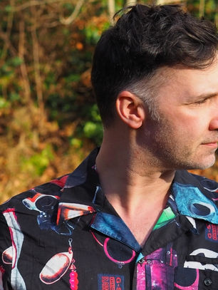 Man in a colorful patterned shirt stands outdoors, looking to the side. Background features lush greenery and a sunny, natural setting.