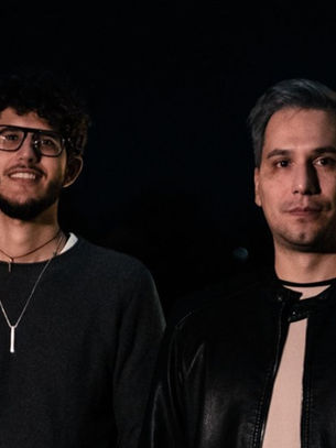 Two men stand side by side at night, one smiling, the other serious. They wear casual dark clothing. Blurred lights glow in background.