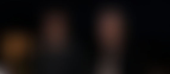 Two men stand side by side at night, one smiling, the other serious. They wear casual dark clothing. Blurred lights glow in background.