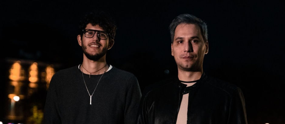 Two men stand side by side at night, one smiling, the other serious. They wear casual dark clothing. Blurred lights glow in background.