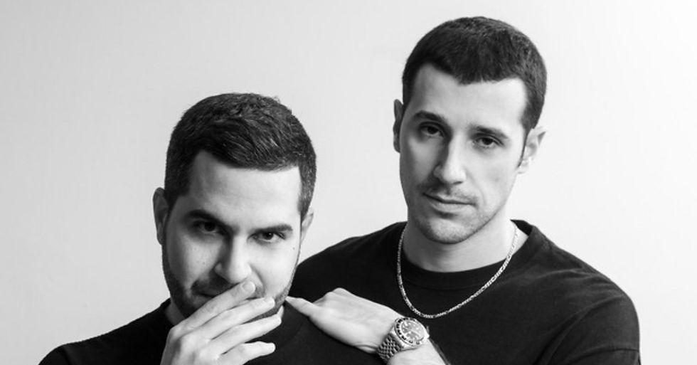 Two men in black shirts pose against a white background. One has his hand near his face, and the other rests his hand on his shoulder. They look calm.
