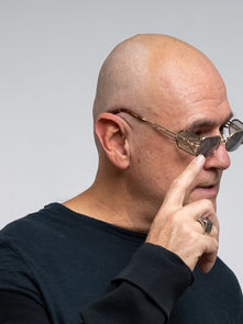 Bald man with glasses and black shirt touches face, looking thoughtful. Neutral background, minimalistic style.
