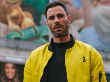 Man in a bright yellow jacket stands in front of a mural with colorful artwork. He has a calm expression. Urban setting.