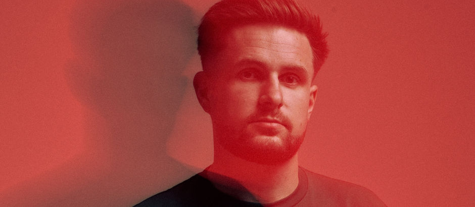 Man with short hair and beard in a red-lit room, casting a shadow on the wall. He looks serious, wearing a dark shirt.