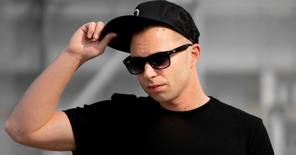 Man in black shirt and cap adjusts his hat. Wearing sunglasses, he stands against a blurred gray background, exuding a casual vibe.