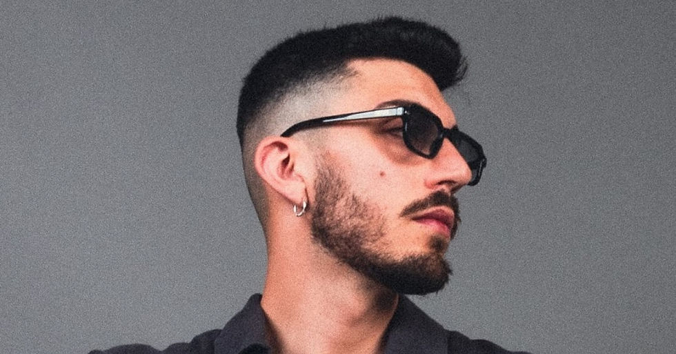 Man with sunglasses and beard, wearing a dark shirt, gazes sideways. He has a short haircut and an earring. Gray background. Mood: relaxed.