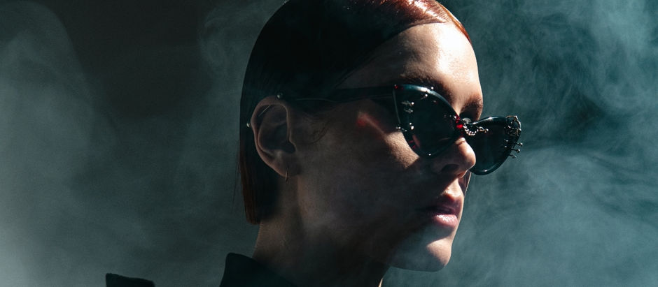 Close-up profile of a woman in black sunglasses and slicked-back hair, lit dramatically in smoky teal light.