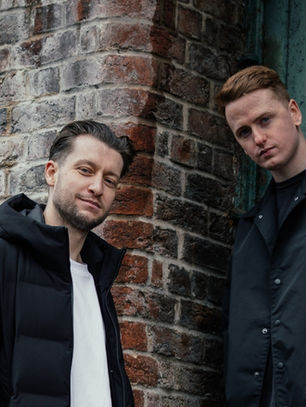 Two men in black jackets lean against a weathered brick wall and teal door, posing with calm, moody expressions.