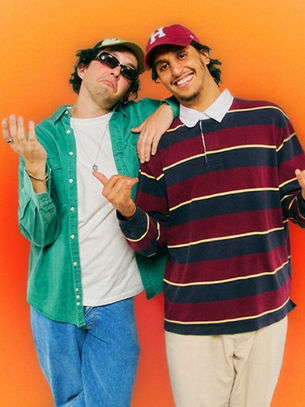 Two smiling people pose against an orange background. One wears a green shirt and cap, the other in a striped shirt and maroon cap. Relaxed vibe.