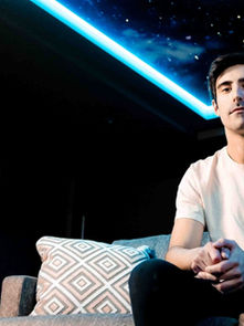 A person sits on a couch with a geometric pillow, under a ceiling featuring blue LED lights and stars. Mood is calm and introspective.