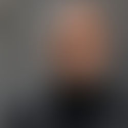 Bald man in a black turtleneck and leather jacket, standing against a concrete wall, looks at the camera with a calm expression.