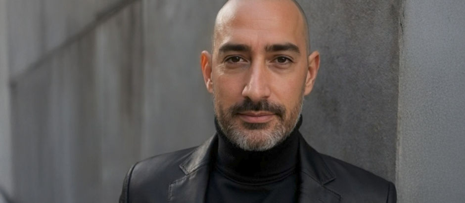 Bald man in a black turtleneck and leather jacket, standing against a concrete wall, looks at the camera with a calm expression.