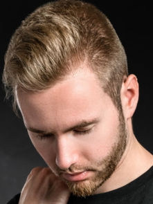 Young man with closed eyes and hand on shoulder, looking down pensively. Dark background, light brown hair, casual black shirt. Emotive mood.