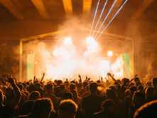 A crowd dances energetically under bright stage lights and smoke at a concert. Colorful lasers pierce the air, creating a vibrant atmosphere.