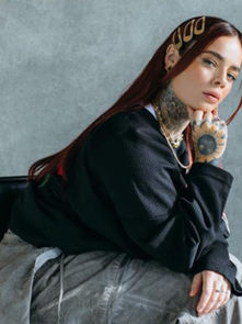 Tattooed woman with red hair sits on a chair, wearing black and gray outfit. Gray background, thoughtful expression, decorative hairpins.