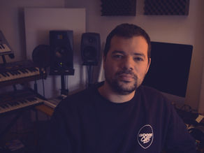Man in a dim home music studio with keyboards, speakers and monitors, wearing a BOOGIE CAFE sweatshirt, looking calmly at camera.