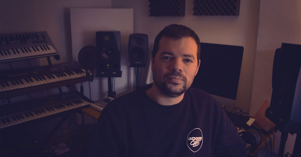 Man in a dim home music studio with keyboards, speakers and monitors, wearing a BOOGIE CAFE sweatshirt, looking calmly at camera.