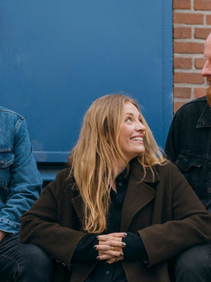 Three people sit smiling against a blue and brick wall backdrop. Two men wear denim jackets, while a woman in a dark coat sits in between.