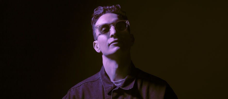 Man in glasses, wearing a dark jacket, lit in purple light against a dark background, appears calm and contemplative.